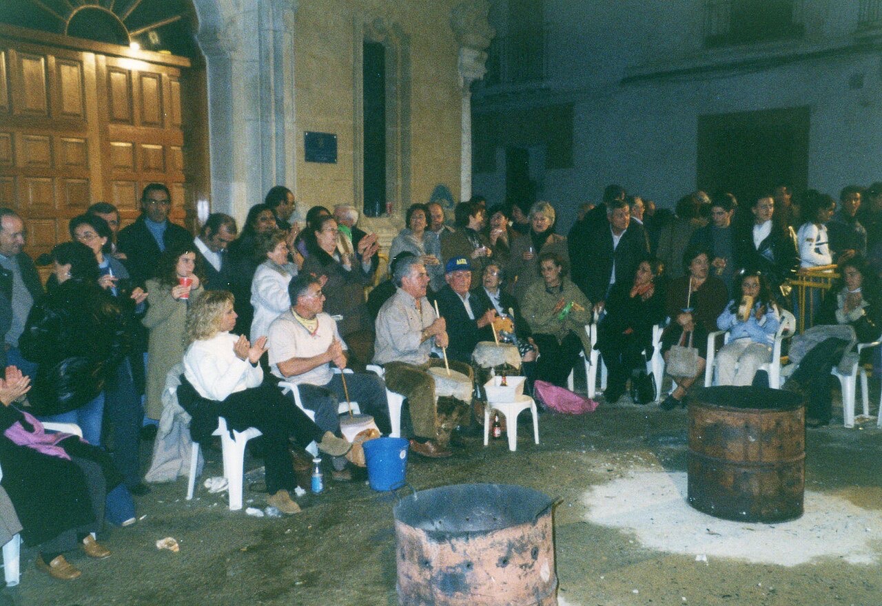 Zambomba de noche en una plaza: vecinos sentados en corro, dos hombres tocando zambombas y dos bidones para la candela.