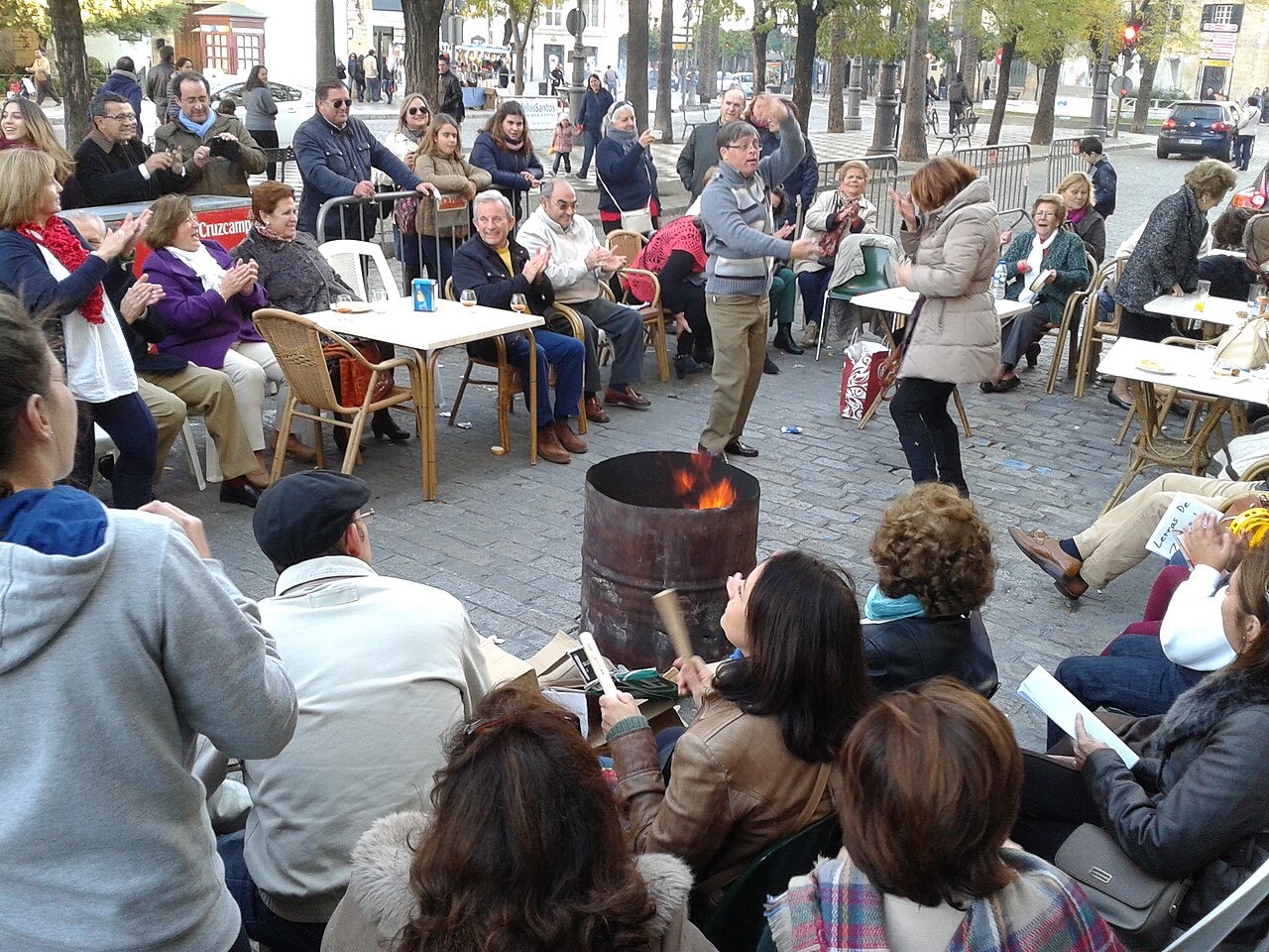 Zambomba en la calle, en la puerta de la capilla de San Juan de Letrán de Jerez: vecinos sentados en corro alrededor de una candela, una pareja bailando en el centro y una mujer tocando la zambomba.