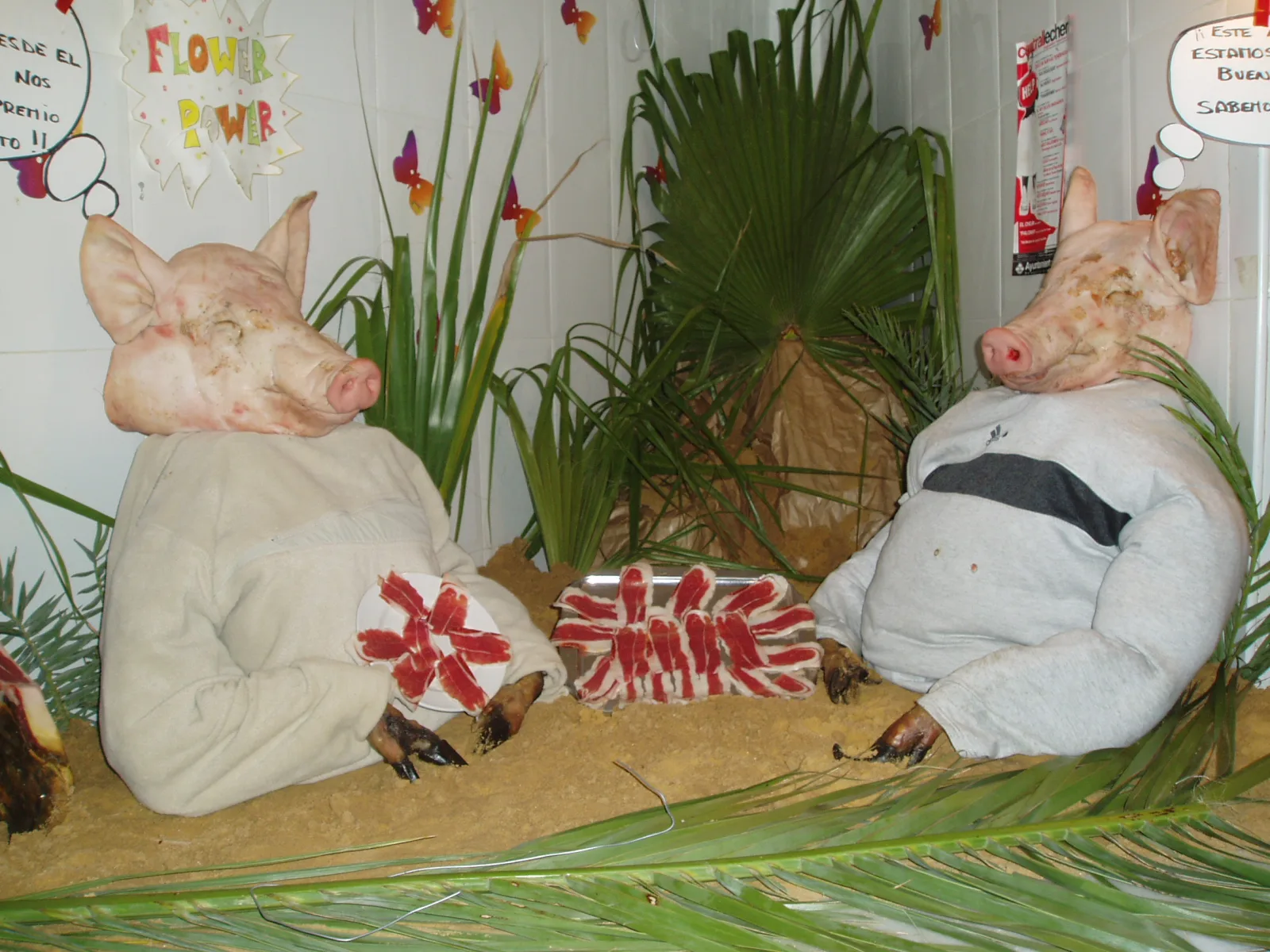 Puesto exornado por Tosantos en un mercado de Cádiz: dos cabezas de cerdo vestidas con ropa blanca entre palmas, con bocadillos de texto humorísticos