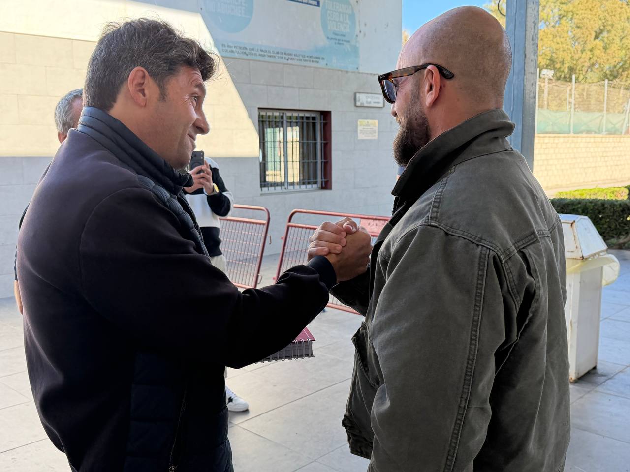 Dos hombres se dan la mano en el exterior de un edificio, con vallas rojas y otra persona al fondo.