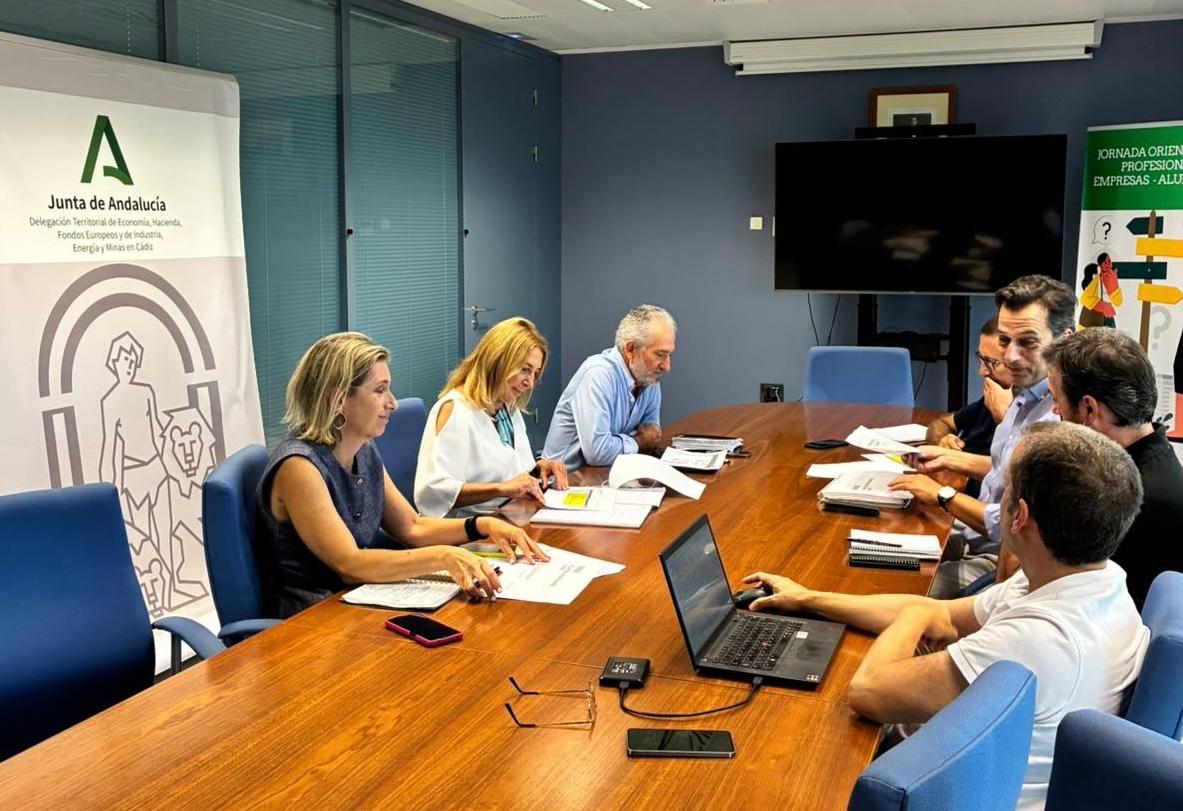 Siete personas sentadas alrededor de una mesa de reuniones con documentos y un portátil, junto a un cartel de la Junta de Andalucía.