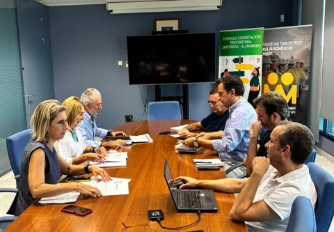 Siete personas sentadas alrededor de una mesa de reuniones con documentos y un portátil, en una sala con pantalla y carteles.