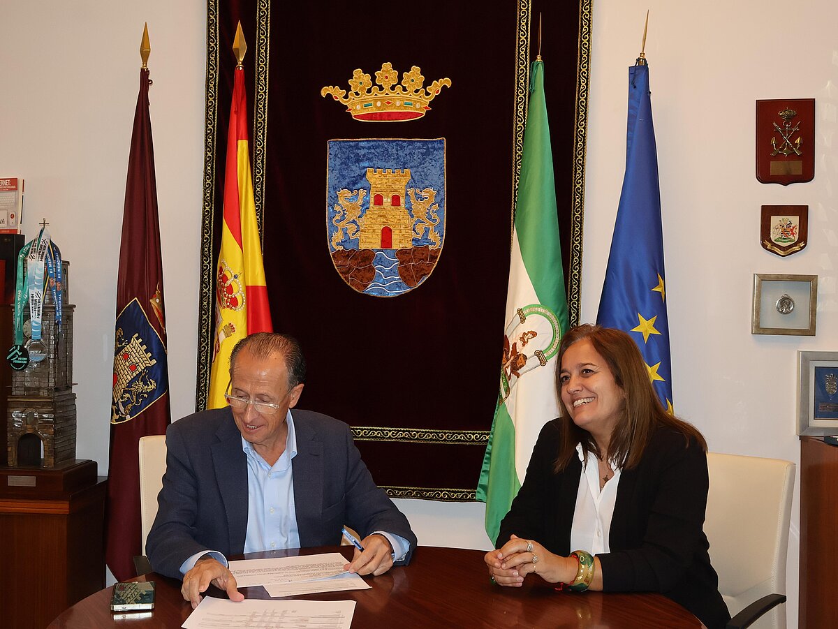 Un hombre con gafas y una mujer sonríen sentados ante una mesa con documentos, con banderas y un escudo bordado al fondo.
