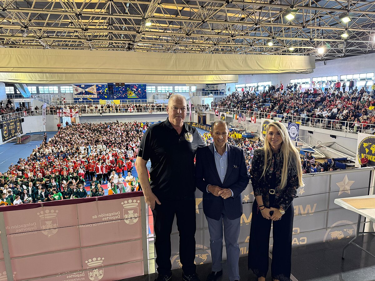 Tres personas de pie tras una barandilla en un pabellón deportivo lleno de público y deportistas en la pista.