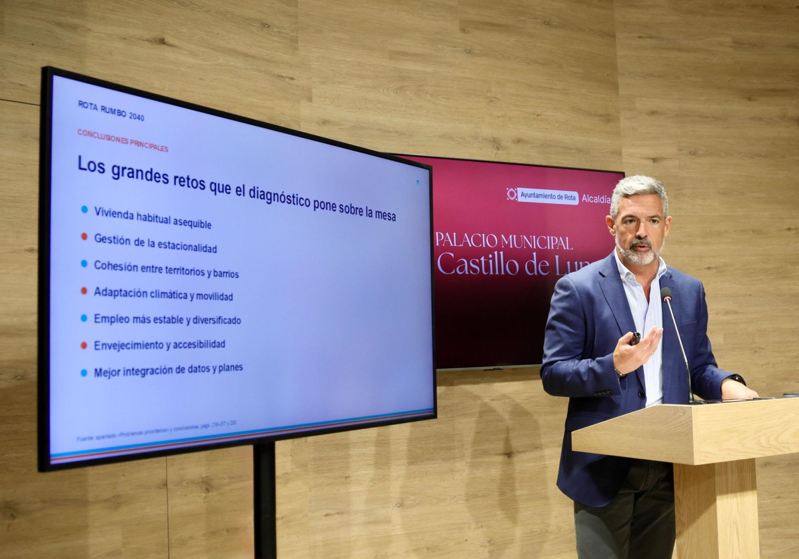 Un hombre con traje azul habla ante un atril junto a dos pantallas con texto sobre retos municipales.