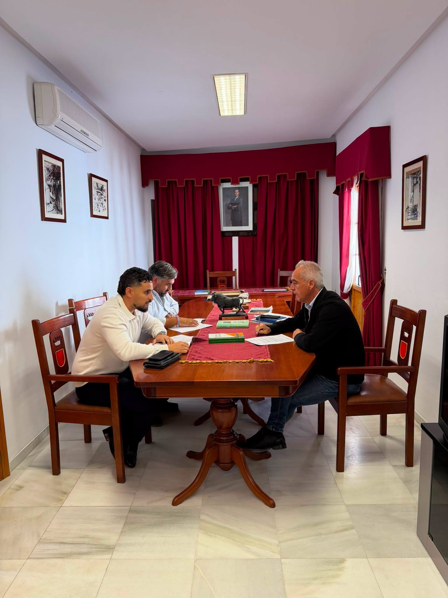 Tres hombres sentados alrededor de una mesa de madera en una sala con cortinas rojas y un retrato en la pared.