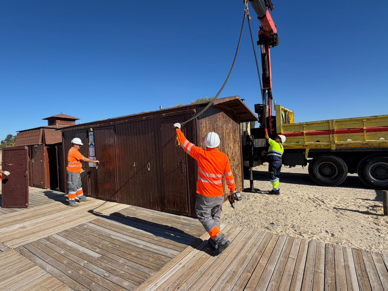 Tres operarios con casco y ropa de alta visibilidad manejan una grúa junto a una caseta de madera sobre una pasarela de tablas en la arena.