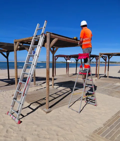 Un operario con casco y chaleco naranja trabaja subido a una escalera sobre una estructura de madera en la arena de una playa.