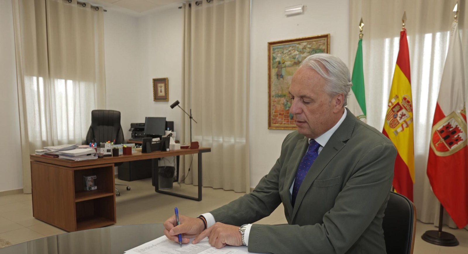 Un hombre mayor con traje gris y corbata azul firma un documento en un despacho con banderas de España, Andalucía y otra local.