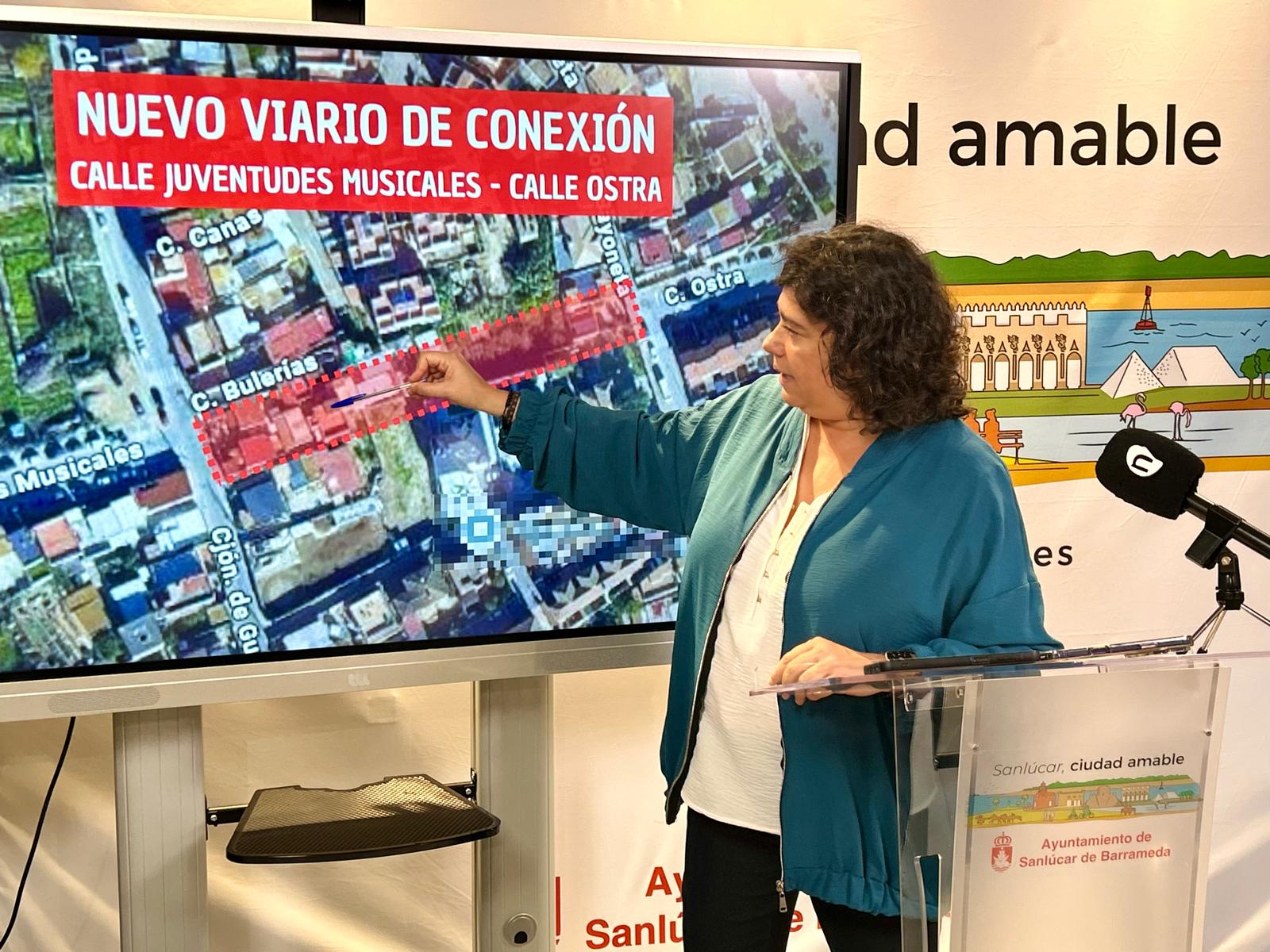 Una mujer señala con un bolígrafo una pantalla que muestra un plano aéreo con calles marcadas en rojo, junto a un atril.