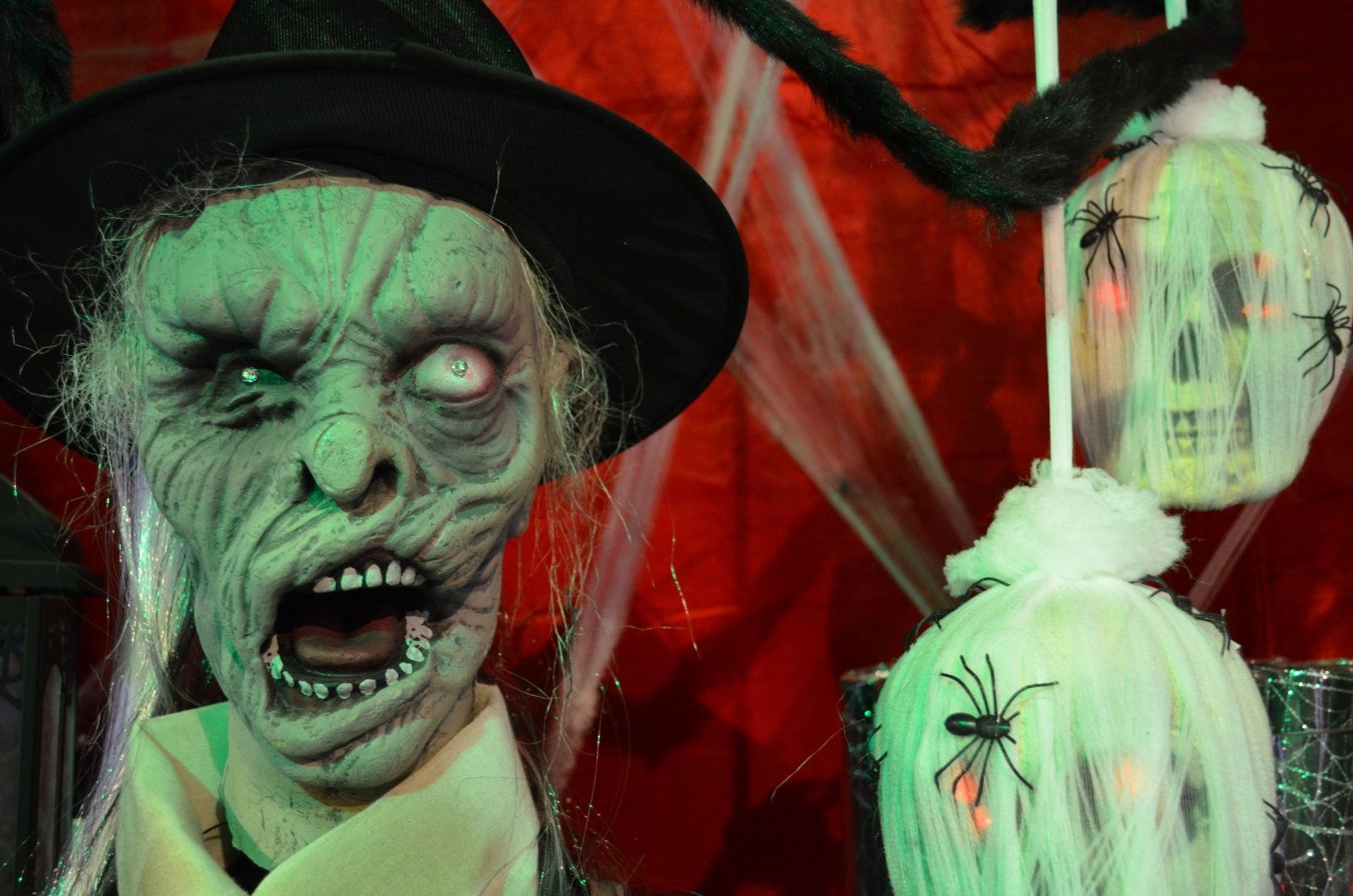 Máscara verde de bruja con sombrero negro y boca abierta, junto a figuras blancas con arañas sobre fondo rojo.
