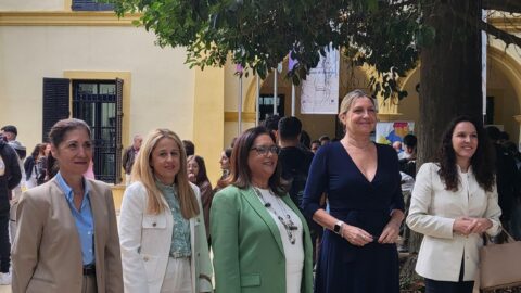 Cinco mujeres con ropa formal posan sonrientes en un patio exterior con edificio amarillo y árboles, con más gente al fondo.
