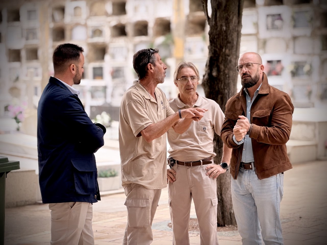 Cuatro hombres de pie conversando en un patio con nichos de cementerio al fondo.
