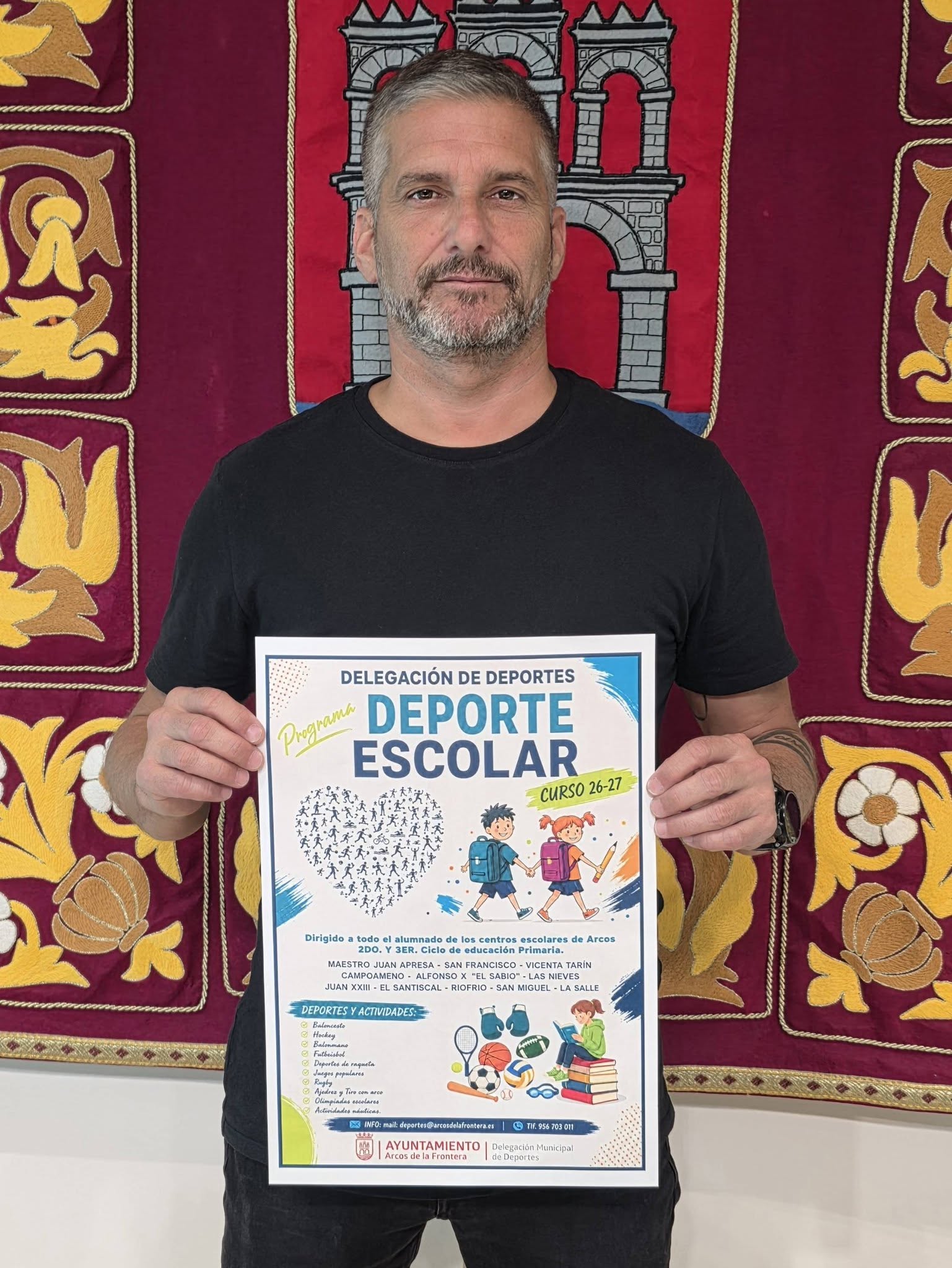 Un hombre con barba y camiseta negra sostiene un cartel de Deporte Escolar ante un tapiz rojo con motivos dorados.