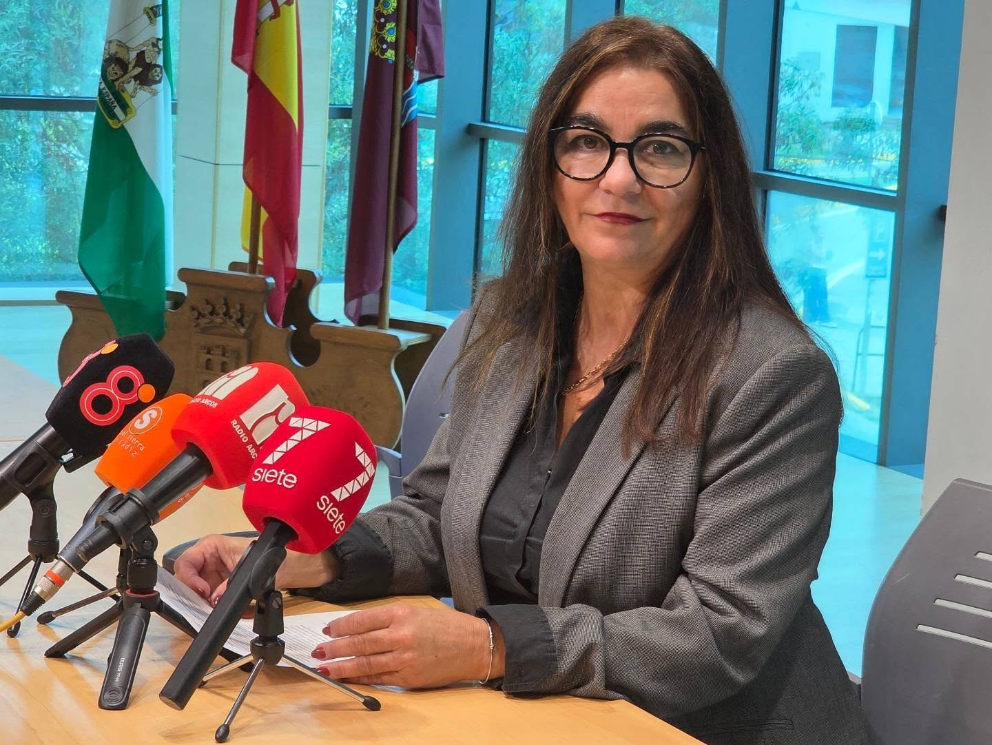Mujer con gafas y chaqueta gris sentada ante una mesa con varios micrófonos de emisoras y banderas al fondo.