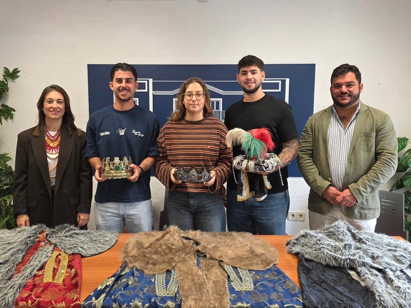 Cinco personas de pie sostienen coronas y un tocado con plumas ante una mesa con ropajes y pieles, con un panel azul al fondo.