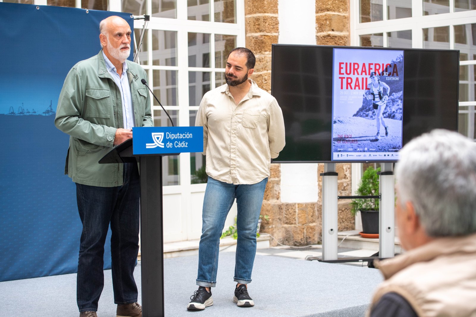 Dos hombres en un estrado, uno habla en un atril con el logotipo de la Diputación de Cádiz y junto a ellos hay una pantalla con un cartel de Eurafrica Trail.