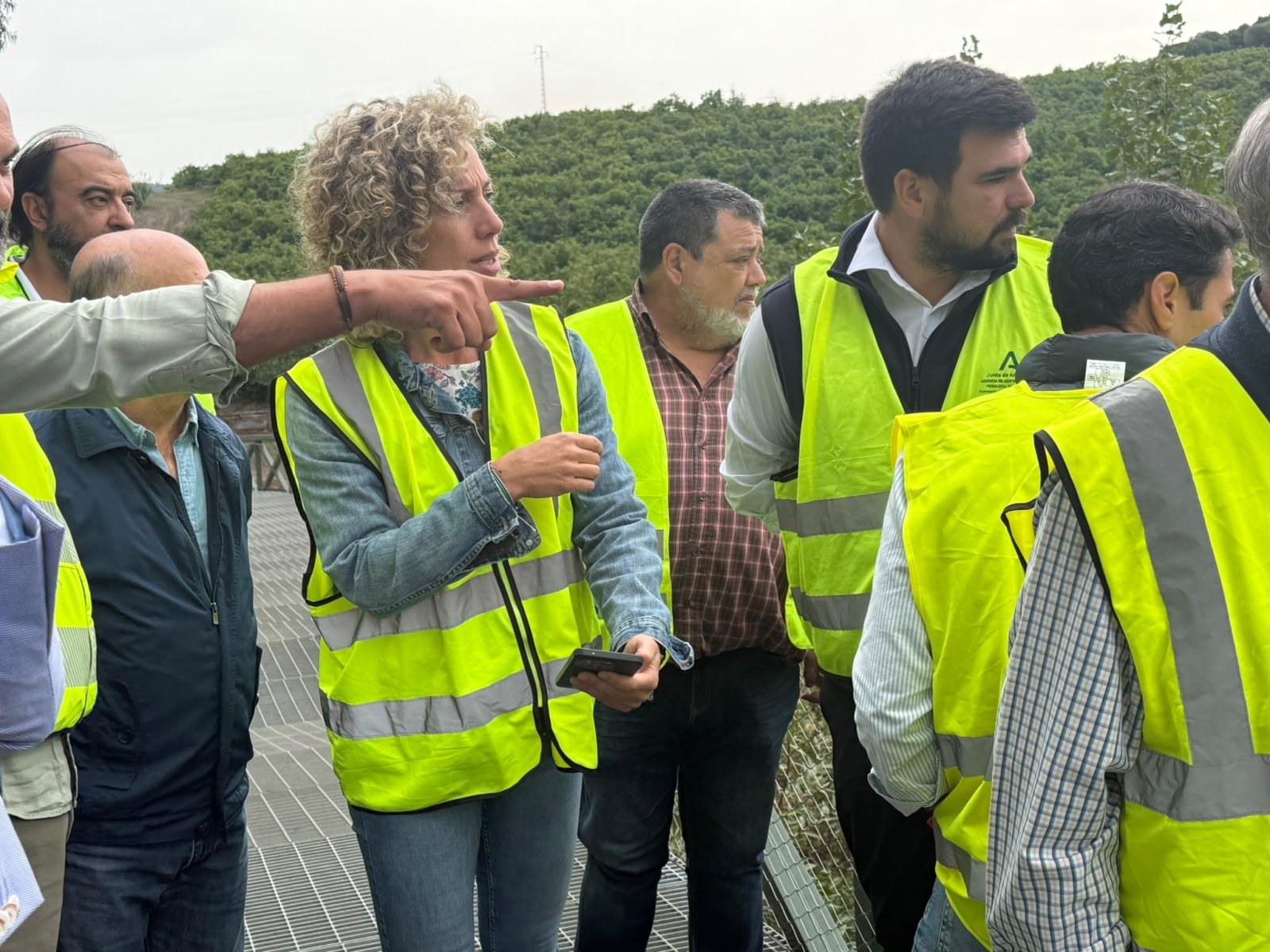 Un grupo de personas con chalecos reflectantes amarillos conversa sobre una pasarela metálica, con colinas verdes al fondo.