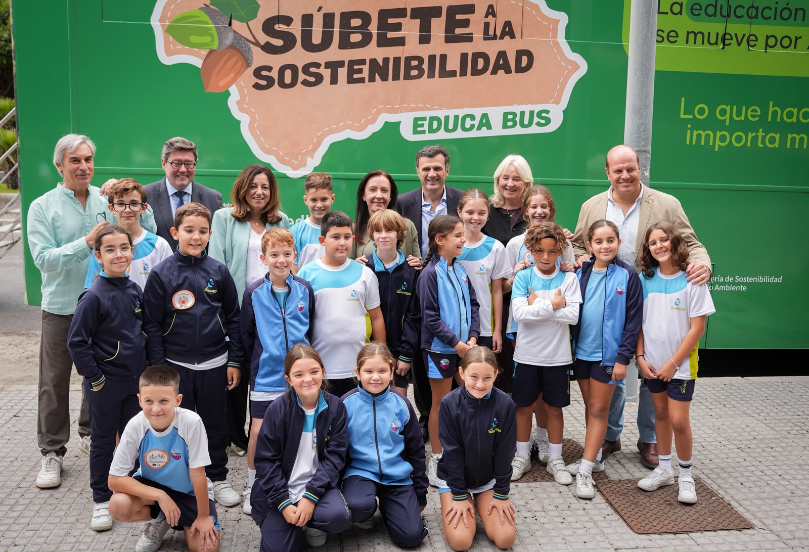 Un grupo de niños con chándal y varios adultos posan sonrientes ante un gran panel verde con el texto Súbete a la Sostenibilidad.
