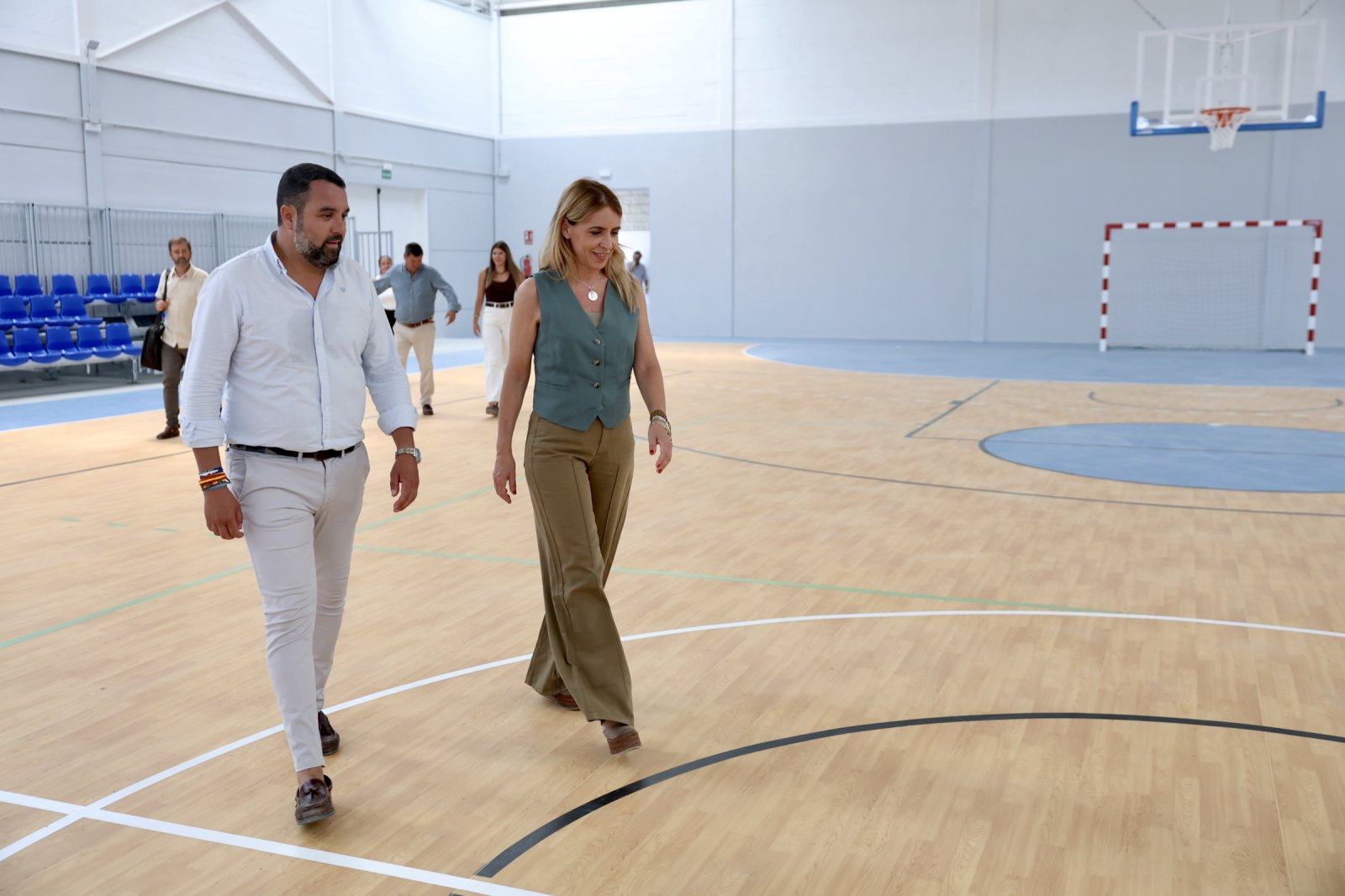 Un hombre y una mujer caminan por la pista de madera de un pabellón deportivo con canasta y portería al fondo.
