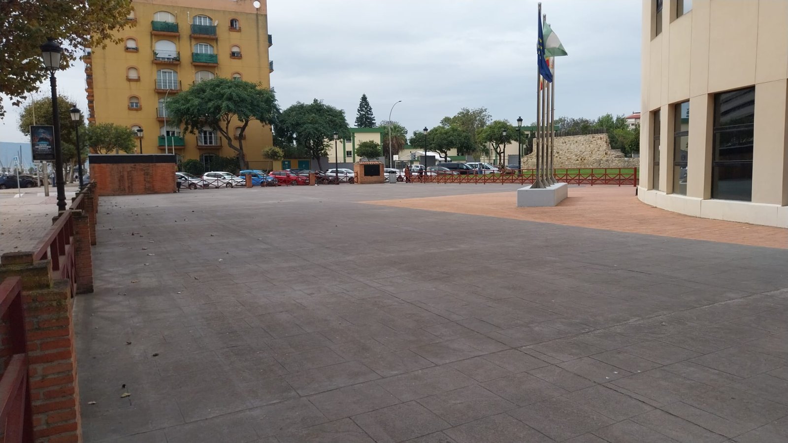 Una plaza amplia y vacía de suelo embaldosado, con un edificio amarillo, coches aparcados, árboles y mástiles con banderas al fondo.