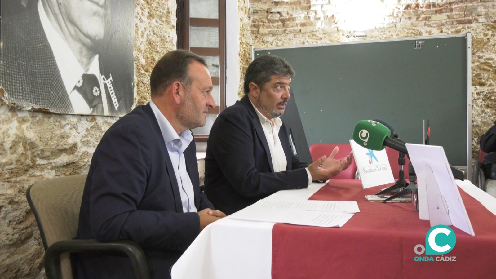 Dos hombres trajeados sentados ante una mesa con mantel rojo y blanco, junto a un micrófono verde y un cartel.