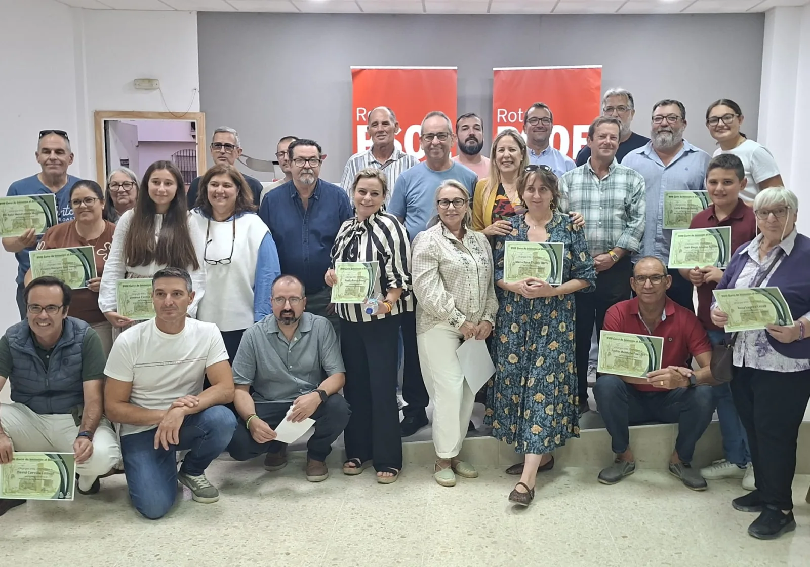 Un grupo de unas veinte personas posa en una sala con diplomas verdes en la mano y carteles rojos al fondo.