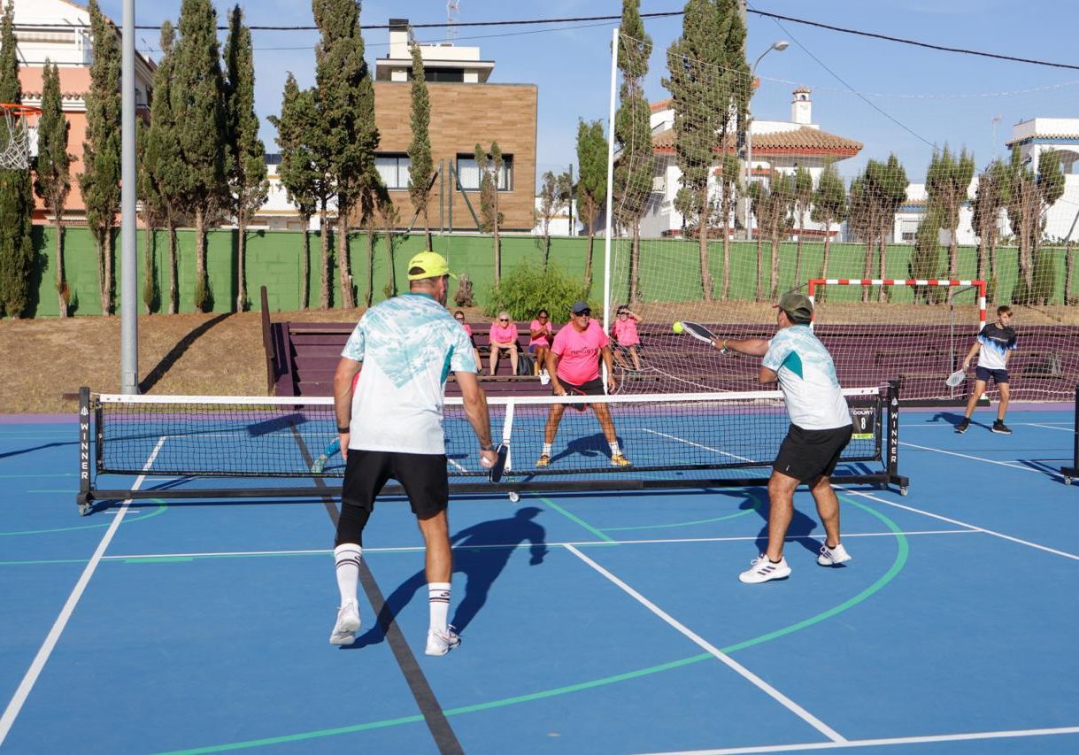 Cuatro jugadores de pádel con palas en una pista azul de exterior, con espectadores sentados al fondo entre árboles y edificios.