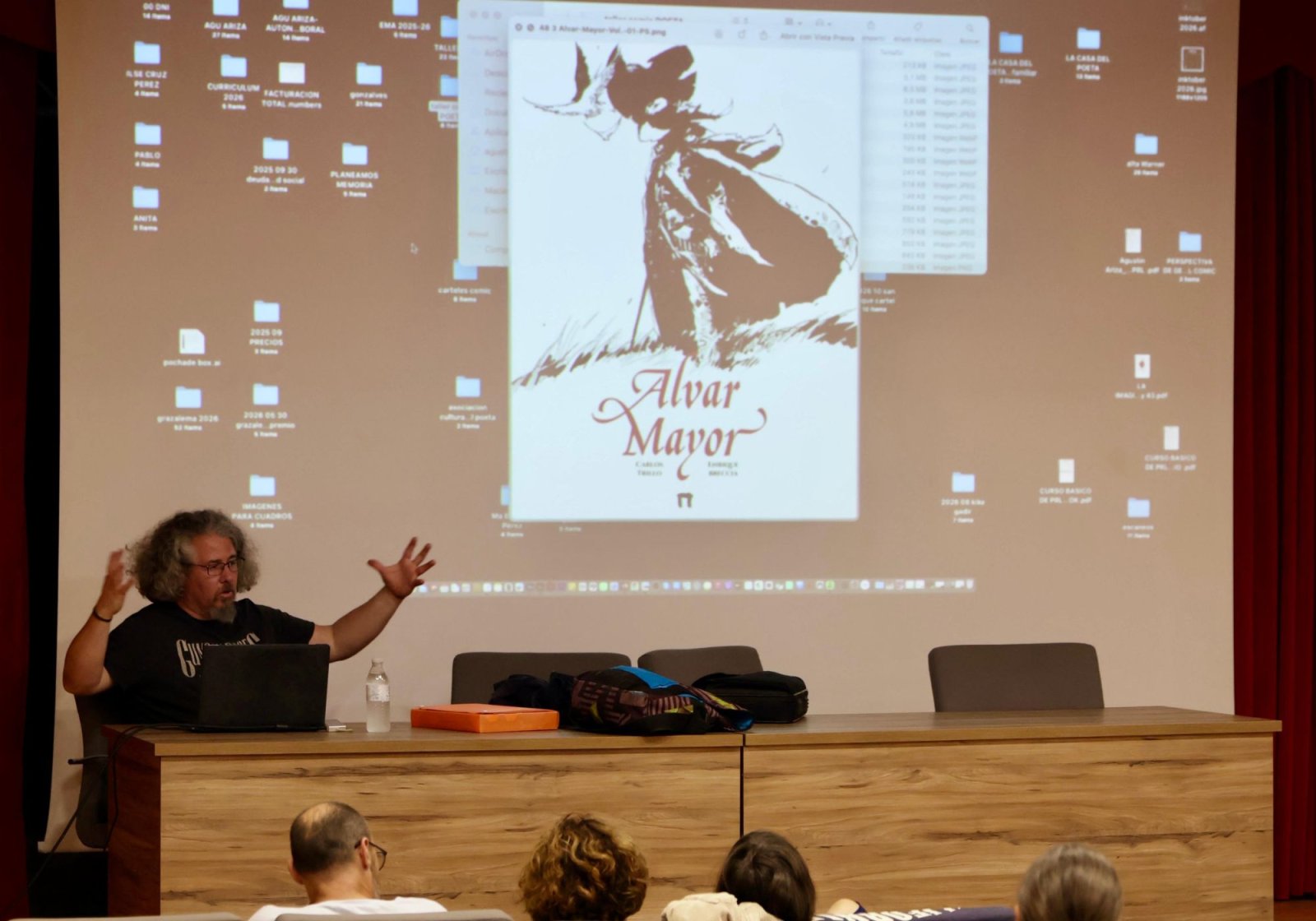 Un hombre con gafas y pelo largo gesticula tras una mesa ante una pantalla que proyecta la portada de un cómic y una carpeta de archivos.
