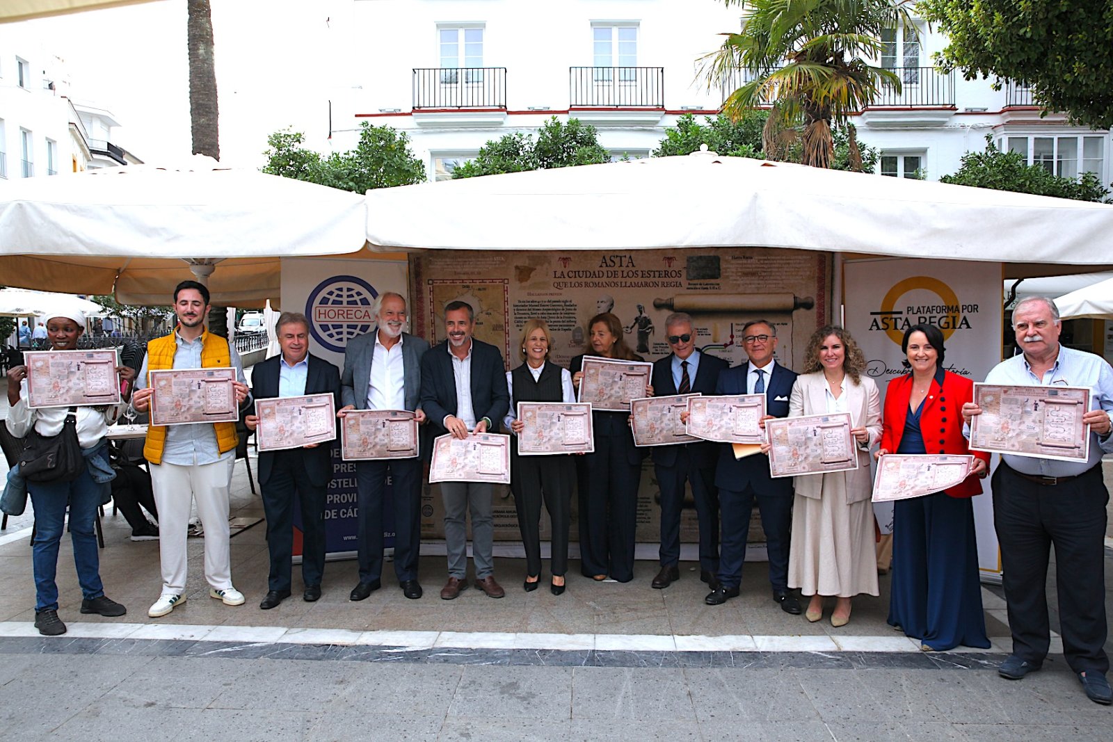 Un grupo de personas posa en la calle sosteniendo carteles con un mapa antiguo bajo una carpa blanca.