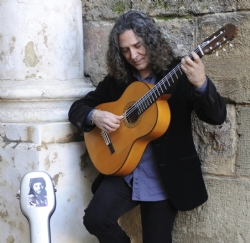 Un hombre de pelo largo y oscuro toca una guitarra española apoyado en un muro de piedra, junto a una funda blanca.