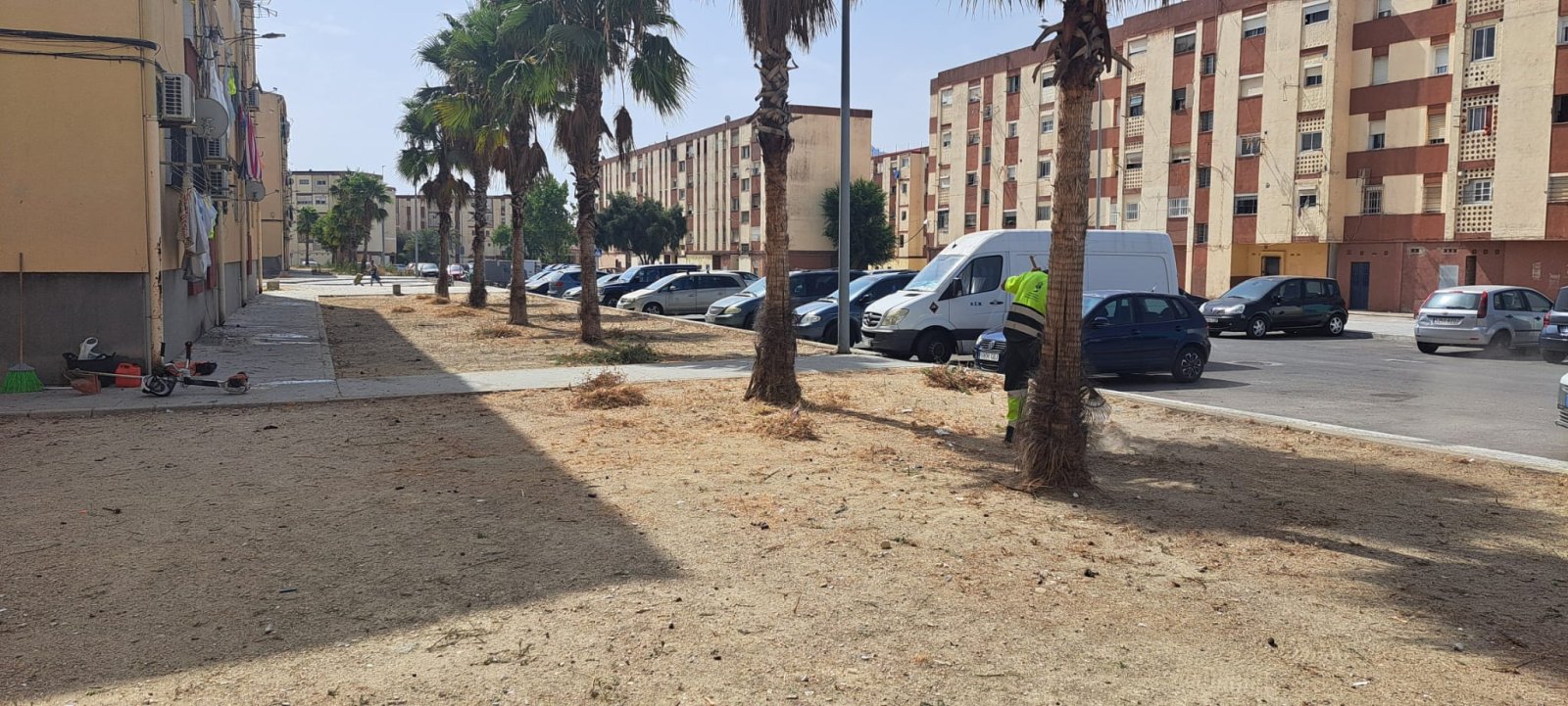 Una persona con chaleco reflectante trabaja junto a una palmera en una zona terriza entre bloques de viviendas y coches aparcados.