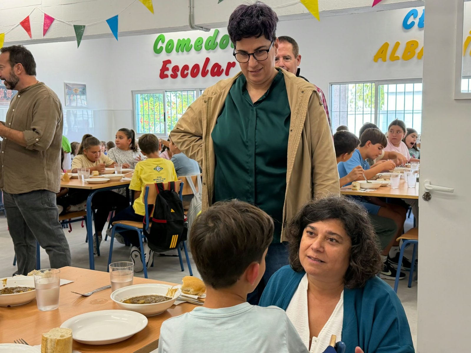 Niños comiendo en un comedor escolar con dos adultos de pie y una mujer sentada junto a un alumno.