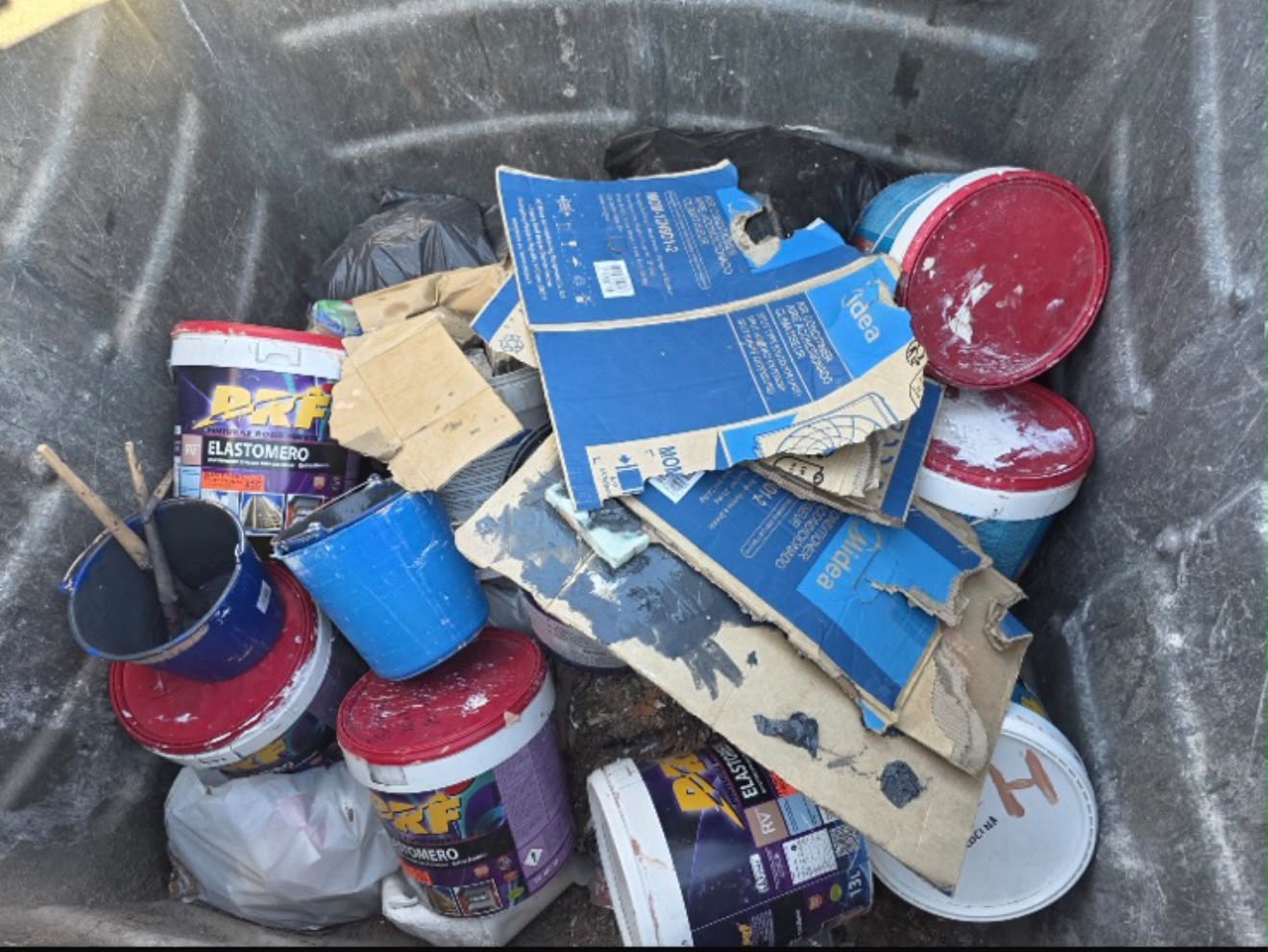 Interior de un contenedor metálico con botes de pintura, cartón y bolsas de basura.