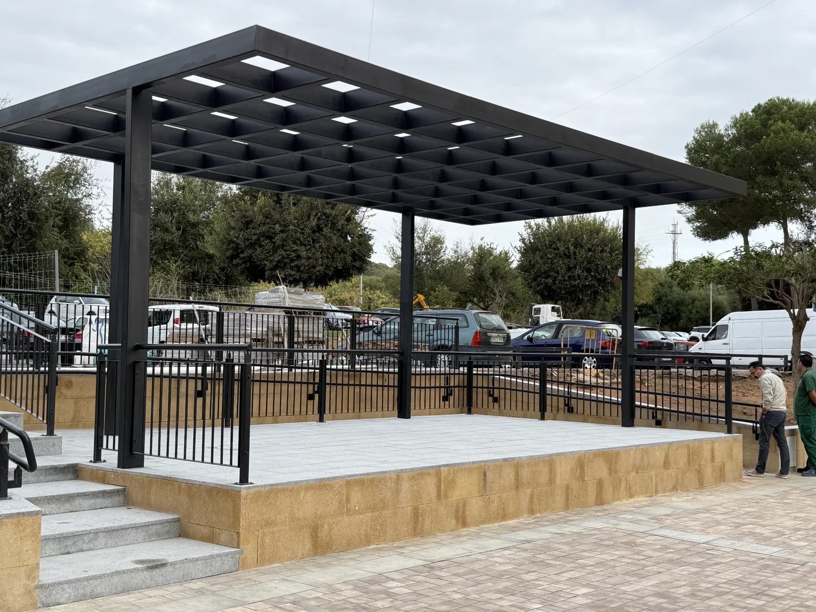 Plataforma elevada de piedra clara con barandilla negra y pérgola metálica oscura con luces, junto a unas escaleras y una zona de aparcamiento.