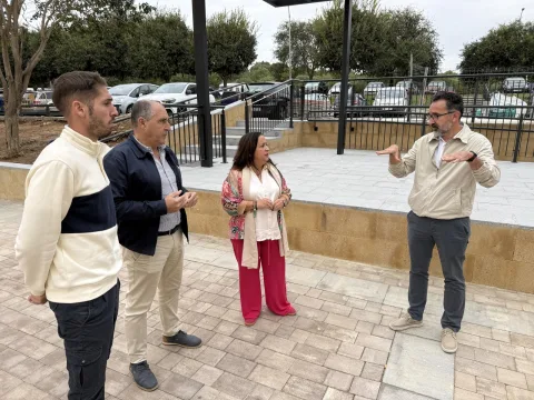 Cuatro personas conversan de pie en una plaza pavimentada junto a una barandilla y coches aparcados al fondo.