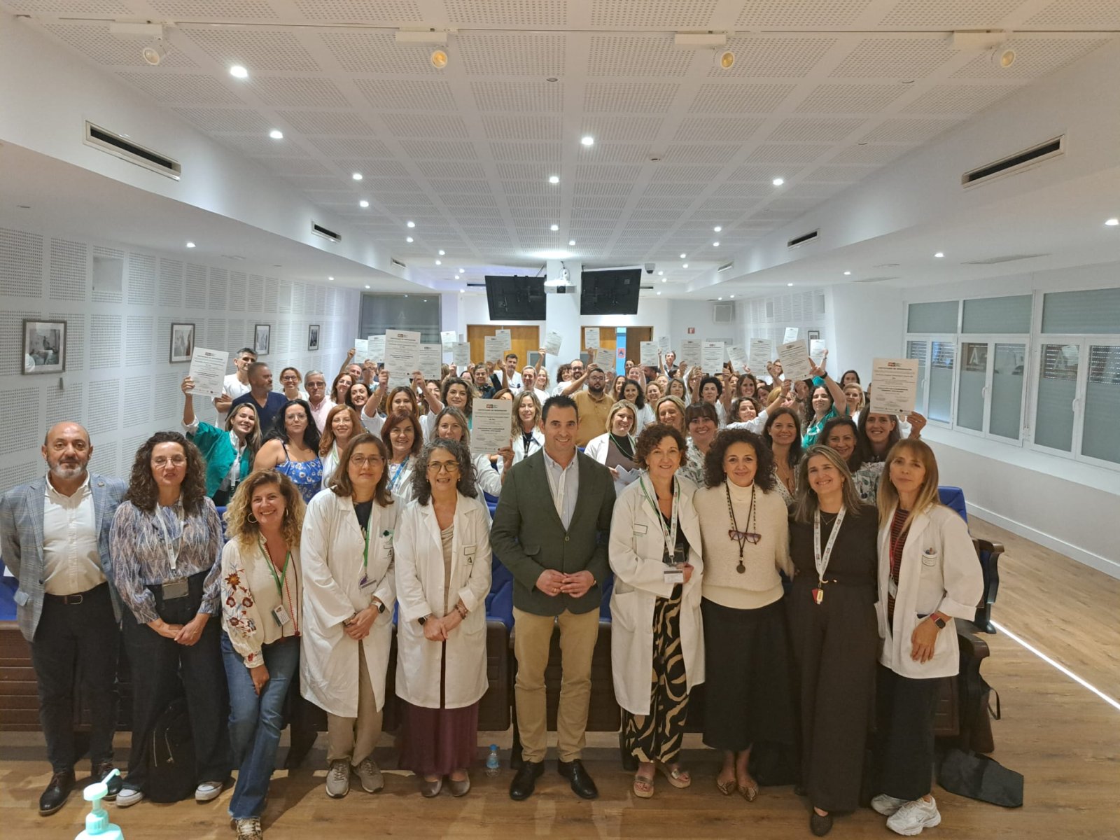 Un grupo numeroso de personas, muchas con bata blanca, posa en una sala amplia con el techo de placas y sostiene documentos en alto.