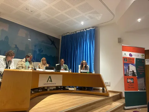 Cinco personas sentadas tras una mesa curva de madera en una sala con cortina azul y un cartel de la Junta de Andalucía.