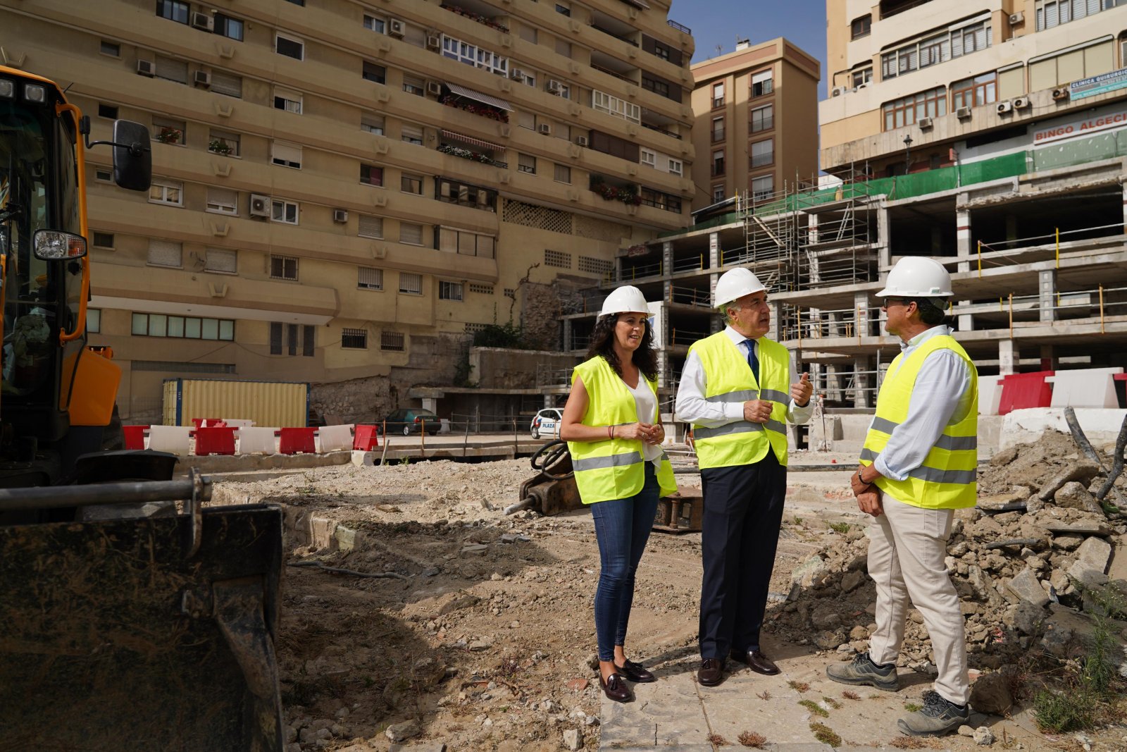 Tres personas con casco y chaleco reflectante conversan en una obra junto a escombros y un edificio en construcción.