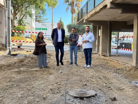 Cuatro personas de pie en una zona de obras con tierra suelta, cinta de balizar y un edificio con aparcamiento al fondo.