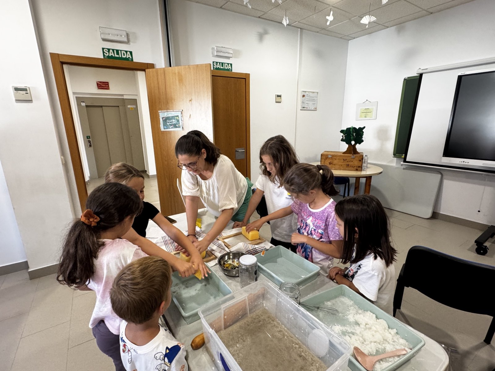 Una mujer adulta y varios niños manipulan esponjas y bandejas con material granular en una mesa dentro de un aula con puertas y carteles de salida.