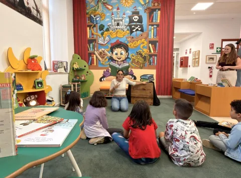 Una mujer sentada en el suelo cuenta un cuento a varios niños en una sala infantil de biblioteca con un gran mural de piratas y libros.