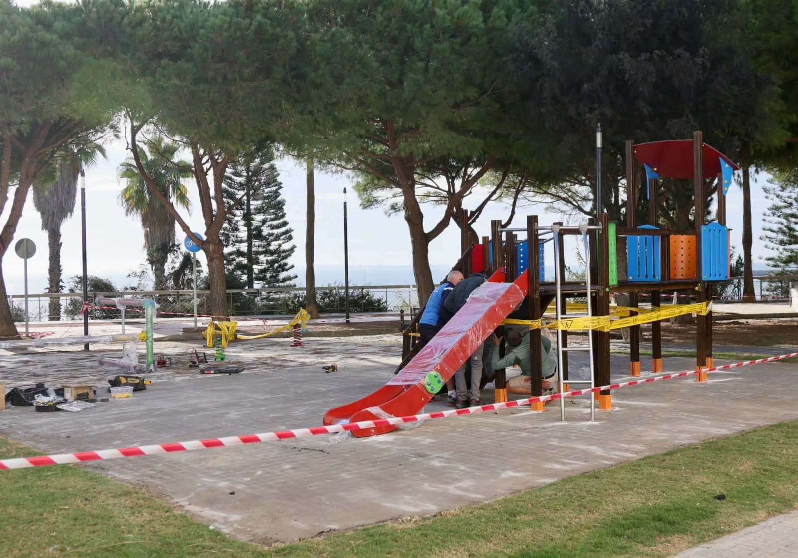 Un parque infantil de colores con tobogán rojo, acordonado con cinta y con operarios trabajando junto a él.
