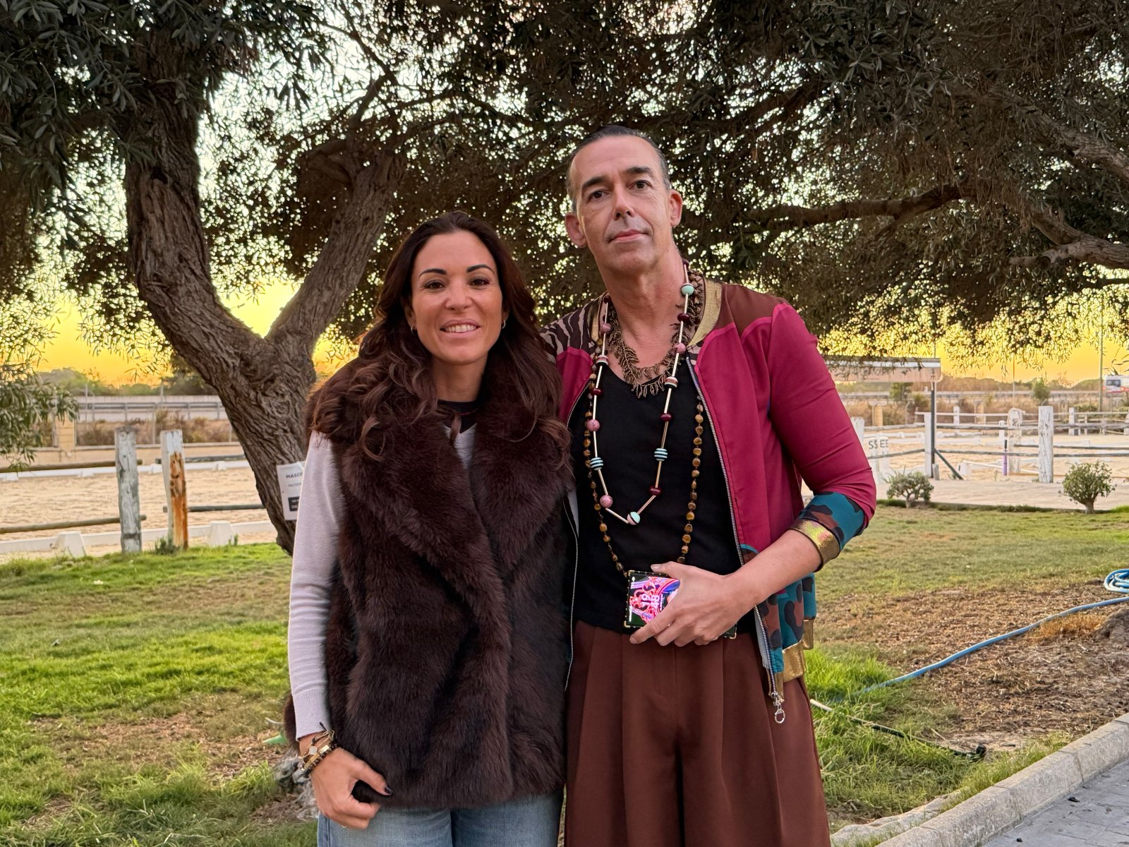Un hombre y una mujer posan sonrientes bajo un árbol, junto a una valla y una pista de arena al atardecer.