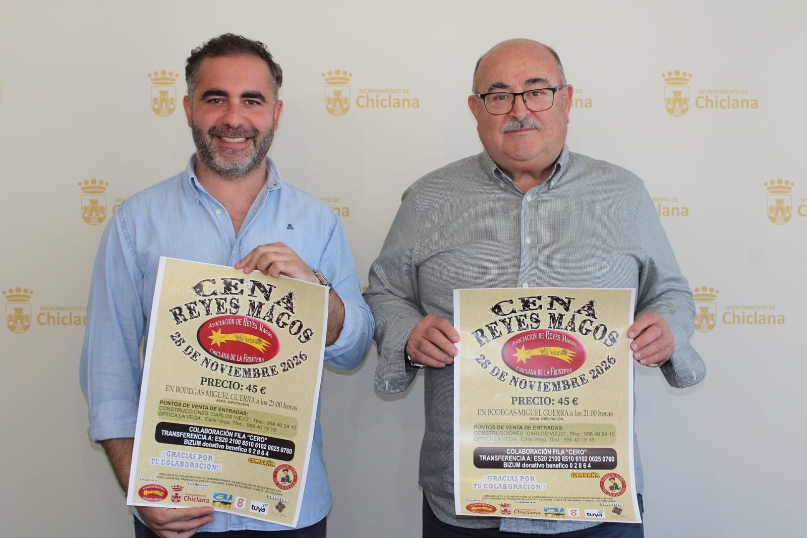 Dos hombres de pie sostienen sendos carteles de la Cena de Reyes Magos ante un fondo con el escudo del Ayuntamiento de Chiclana.