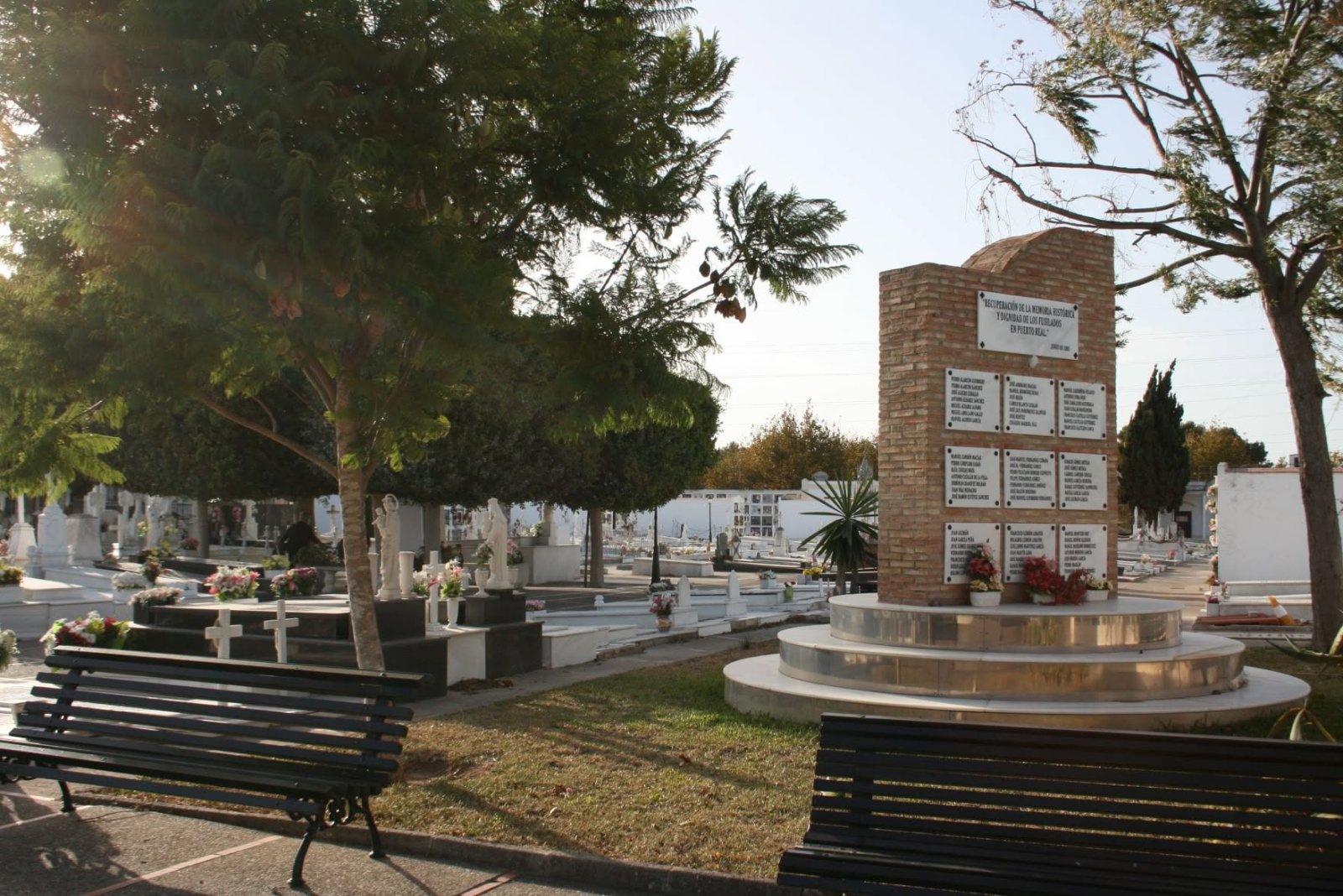 Un monumento de ladrillo con placas conmemorativas en un cementerio, con tumbas, árboles y bancos alrededor.
