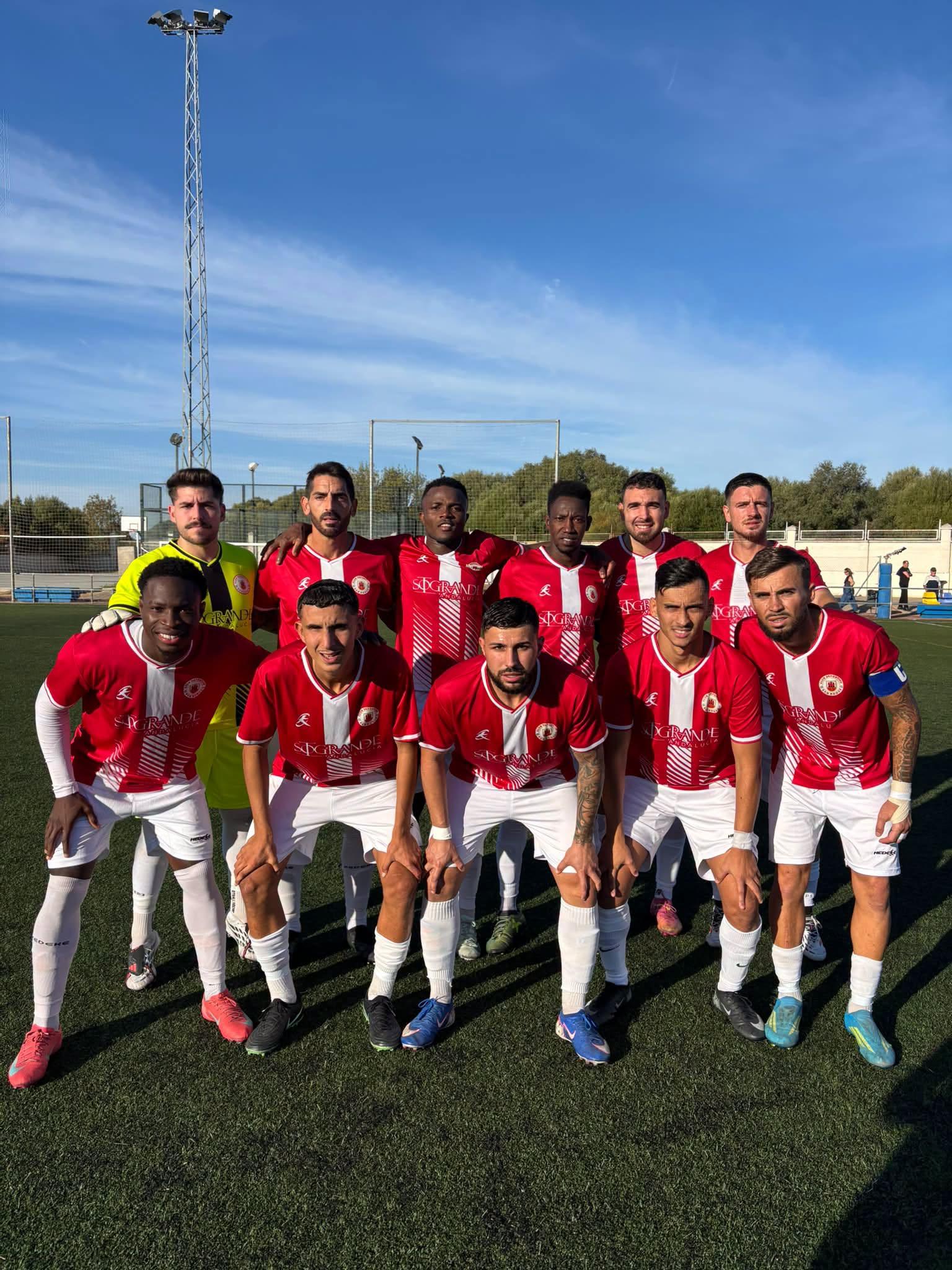 Once jugadores de fútbol con camiseta roja y blanca posan en fila sobre césped, con un portero de verde y un poste de luz al fondo.