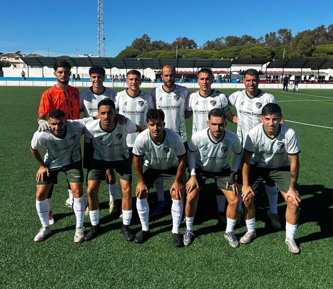 Once jugadores de fútbol posan en dos filas sobre césped verde, con equipación blanquiverde y un portero de naranja, ante gradas y árboles al fondo.