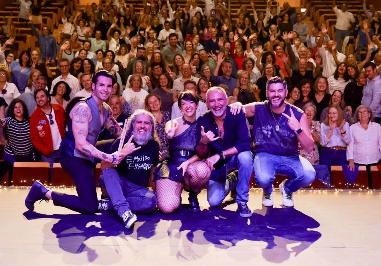Cinco artistas posan en primer plano ante un auditorio lleno de público con los brazos levantados.