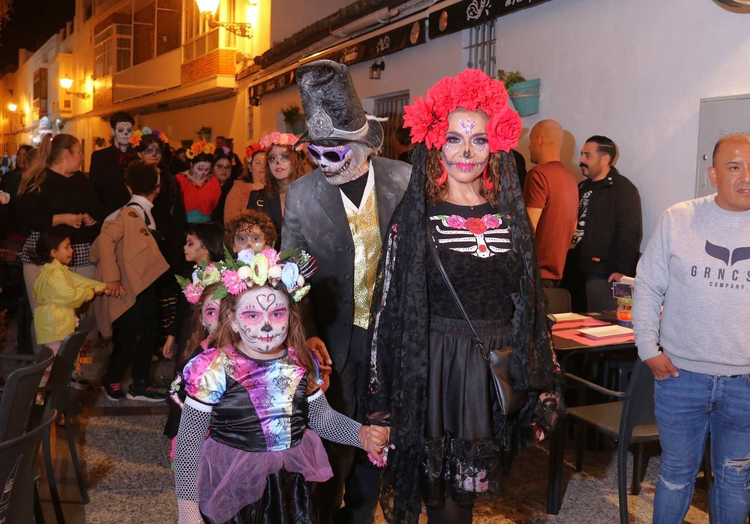 Desfile nocturno en una calle con adultos y niños disfrazados de catrinas y calaveras, con maquillaje y flores en el pelo.